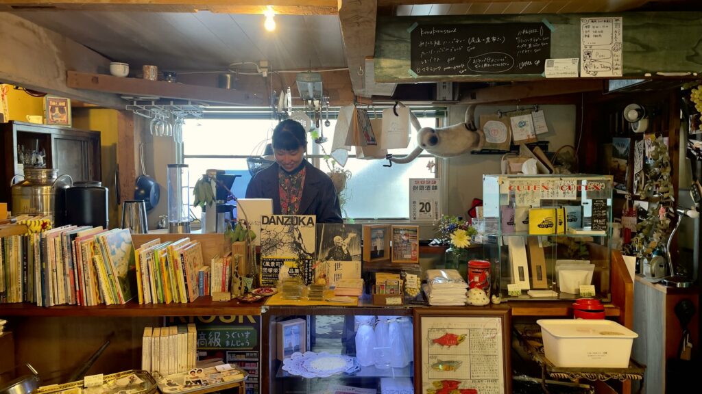 Innenansicht des KURUKURU mit Büchern und Kunstgegenständen, der Getränkekarte. Hinter dem Tresen steht die Betreiberin Miki.
